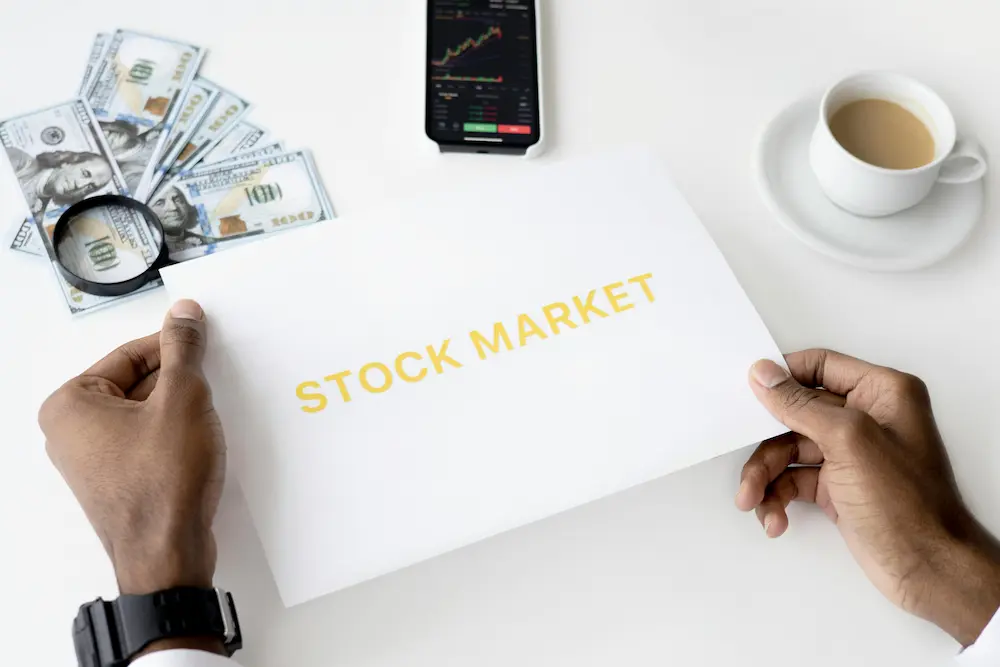 Persona revisando documento con la palabra 'Stock Market', junto a dinero en efectivo, móvil con gráfico de trading y café, representando el entorno del trader moderno.